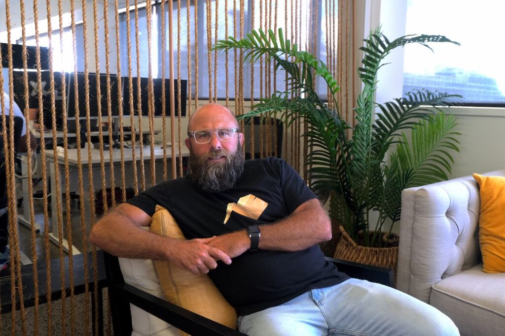 Electric Kiwi CEO, wearing eyeglasses and sitting on a chair in the Electric Kiwi office.